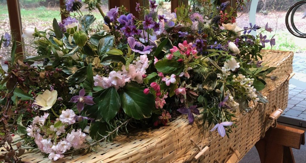 Wicker coffin topped with seasonal wildflower arrangement at a natural burial ceremony